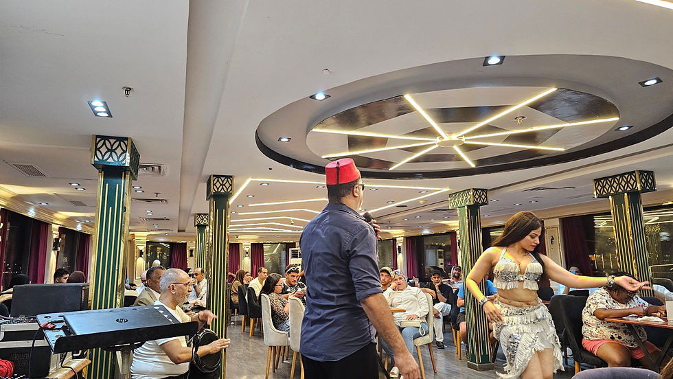 A man with a red hat speaks into a mic as a dancer performs in a lively restaurant with ornate columns. Patrons watch attentively.