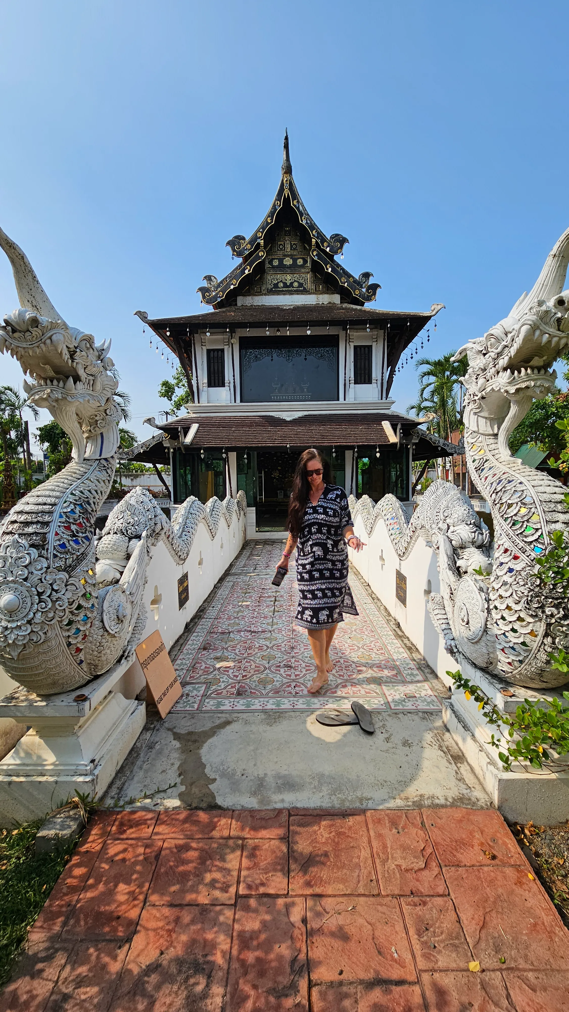 A person in a striped dress walks across a decorative bridge flanked by white dragon statues toward a traditional, ornate building with a tiered roof under a blue sky.