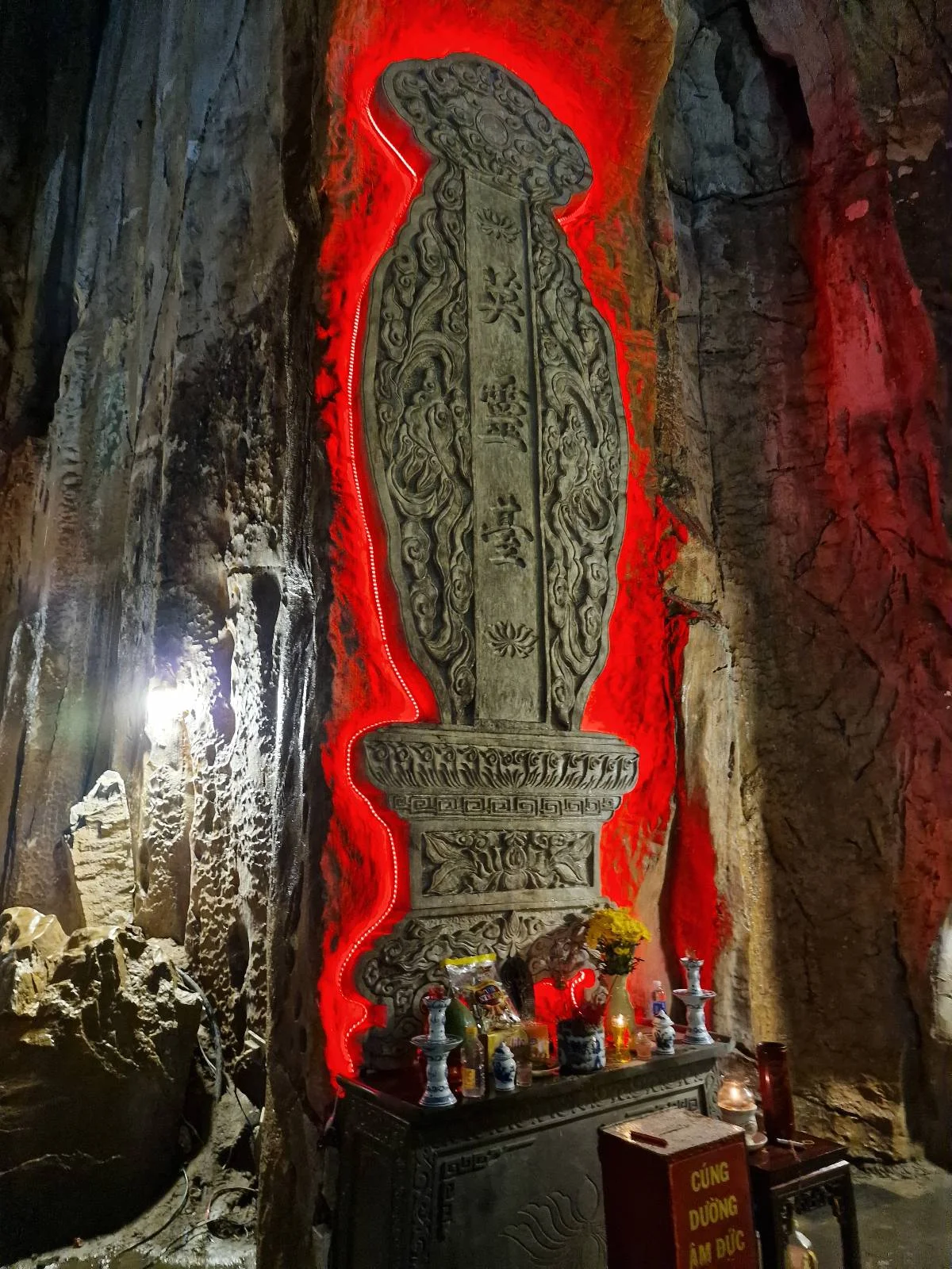 An ornate stone pillar with intricate carvings is illuminated by red light in a cave setting, surrounded by offerings such as flowers and small vases.