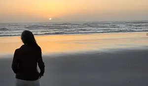A person with long hair stands on Daytona Beach at sunset, facing the ocean. The sky glows orange and yellow above gentle waves, perfect for oceanfront strolls as reflections shimmer across the wet sand.