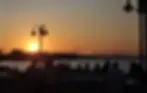 Silhouetted group dining outdoors at sunset by the water, with sailboats in the background. Warm orange and gold hues create a peaceful mood.