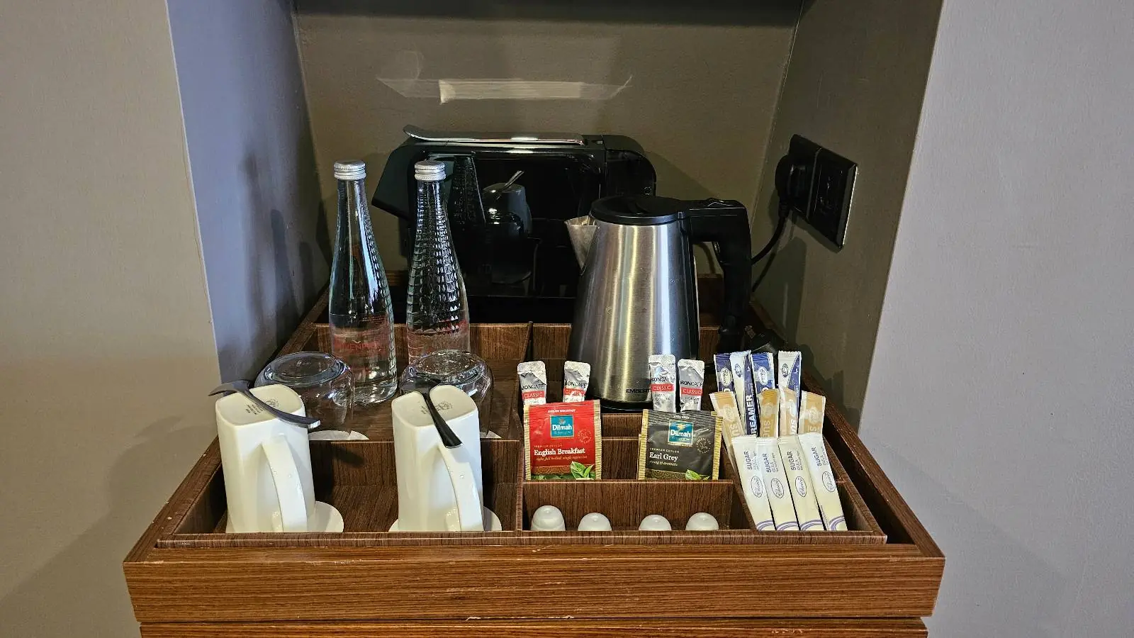 A hotel room refreshment tray featuring a kettle, two glass water bottles, and an assortment of tea and coffee packets. The setup includes sugar, creamer, and stirrers, neatly organized in a wooden compartment.