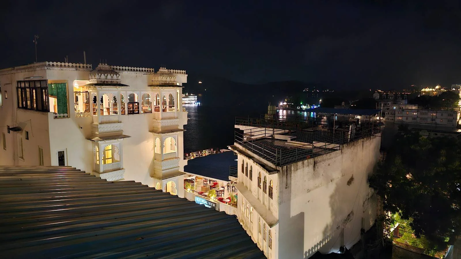 Night view of a white building with balconies and lit windows, overlooking a calm body of water with reflections and distant lights in the background.