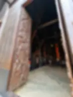 An intricately carved wooden door is partially open, revealing a glimpse inside a dimly lit, spacious room with people walking around.