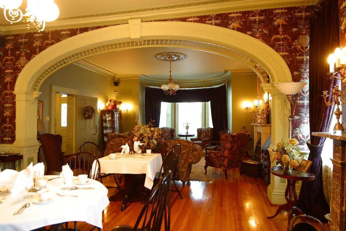 A cozy vintage-style dining room with wooden floors, ornate archways, and dim lighting. Tables are set with white tablecloths and chairs. The decor includes wallpaper, chandeliers, and plush seating in the background.