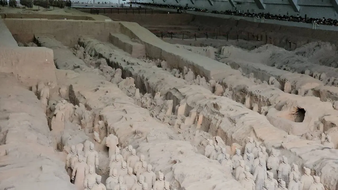Rows of ancient terracotta warrior statues partially buried in an archaeological excavation site, covered by a sheltering roof structure above.