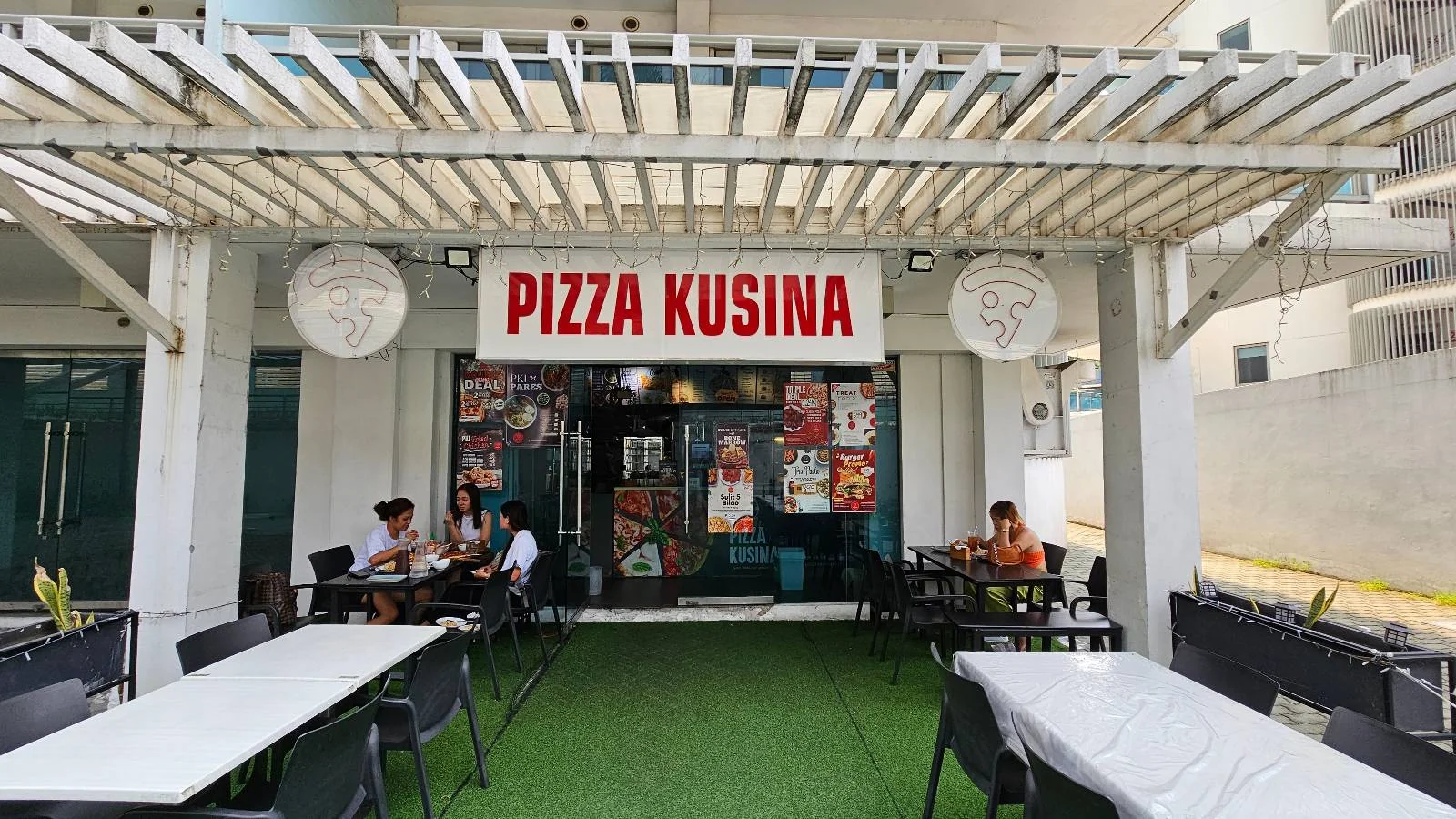 Outdoor view of a restaurant called "Pizza Kusina" with a white pergola, green artificial grass, white tables, and a few people seated and dining outside the entrance.