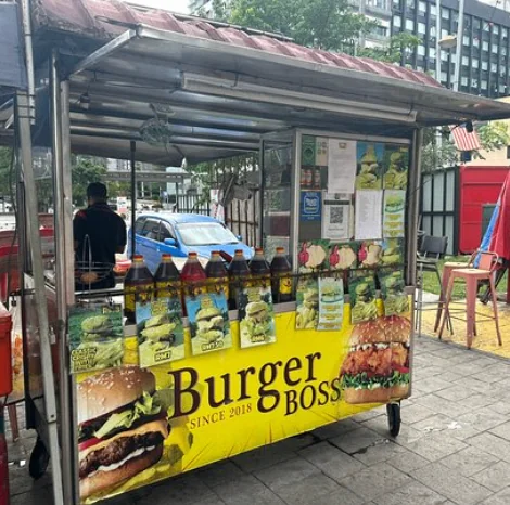 A street food stall named "Burger Boss," adorned with images of burgers and promotional materials. Bottles filled with sauce are lined up on the counter. A person wearing a cap stands inside, ready to serve. The ambiance is vibrant and inviting.