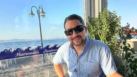 Leon seated at Mavromatis Taverna in Greece, wearing sunglasses with the sea, beach seating, and restaurant signage visible in the background.