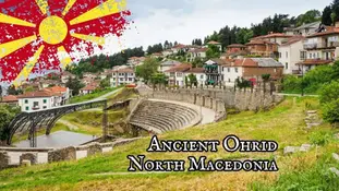 View of the ancient theater in Ohrid, North Macedonia, a true Balkan gem surrounded by lush greenery and traditional houses on a hillside. With Lake Ohrid nearby, the Macedonian flag is stylized in the top left corner. Text reads "Ancient Ohrid North Macedonia.