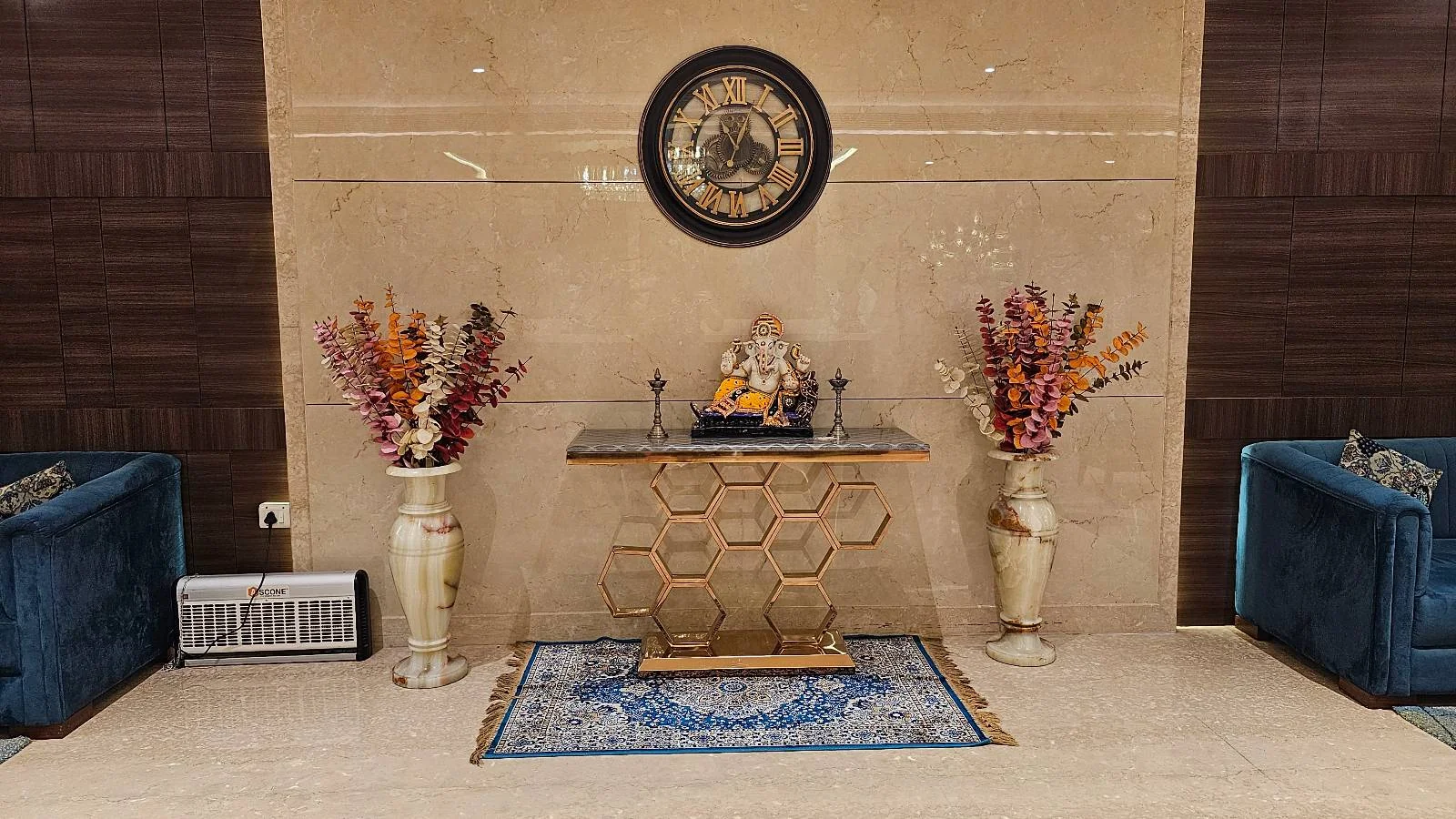 A decorative table with a Ganesha idol stands against a beige wall, flanked by two tall vases with colorful dried flowers. A round wall art piece hangs above, and two blue chairs are placed on either side.