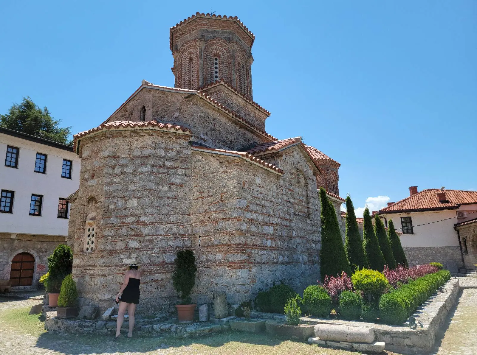 A person in black clothing stands near a historic stone church with a tall, cylindrical tower and red-tiled roof. The church is surrounded by trimmed hedges and trees under a clear blue sky.