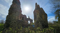 Stone ruins stand tall against a partly cloudy sky with the sun shining brightly above. Surrounding vegetation includes trees and bushes.