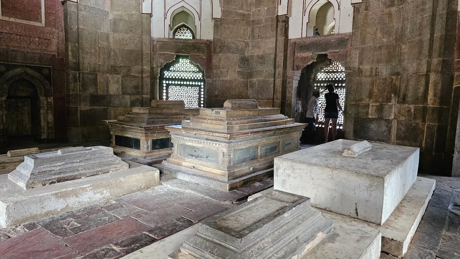 Several stone tombs and sarcophagi are arranged inside a historic building with arched doorways and windows. Two people are visible in the background, standing near the entrance.