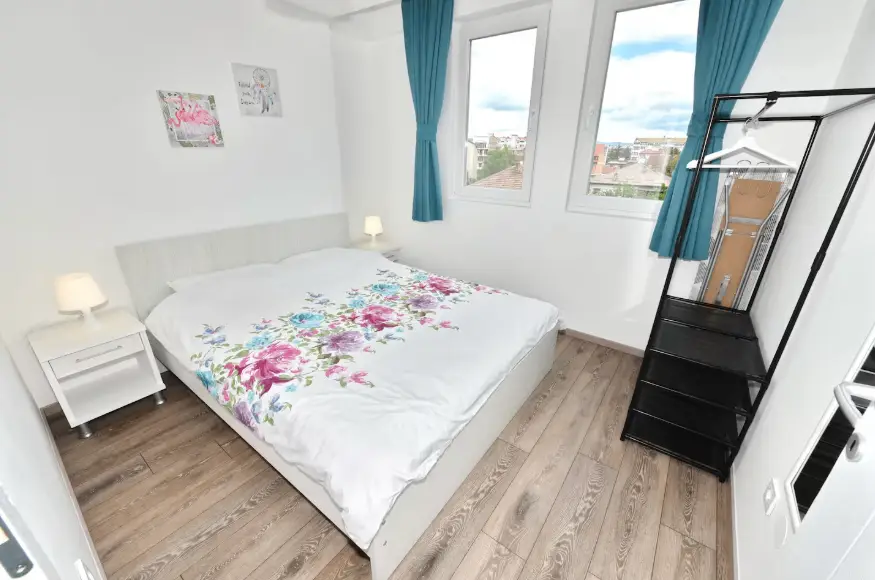 Bright bedroom with a double bed, floral bedding, two nightstands with lamps, wall art, blue curtains, two windows, and a black open wardrobe on a light wood floor.