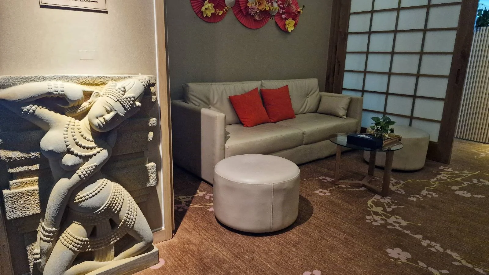 A modern living room with a beige sofa and two red cushions. A circular ottoman and small table with a plant are on a floral carpet. To the left, there's an intricately carved stone sculpture on the wall.