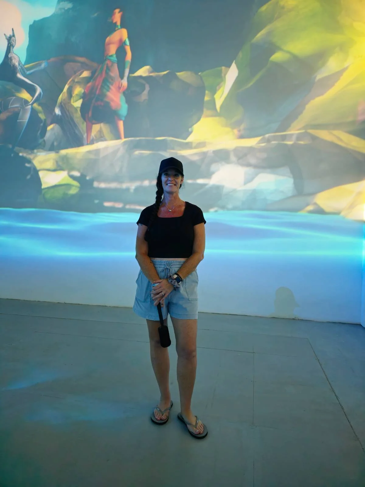 A person in a black shirt and denim shorts stands in an art gallery with vibrant, colorful digital art projected on the wall behind them. The scene includes a person in a red outfit and abstract elements.