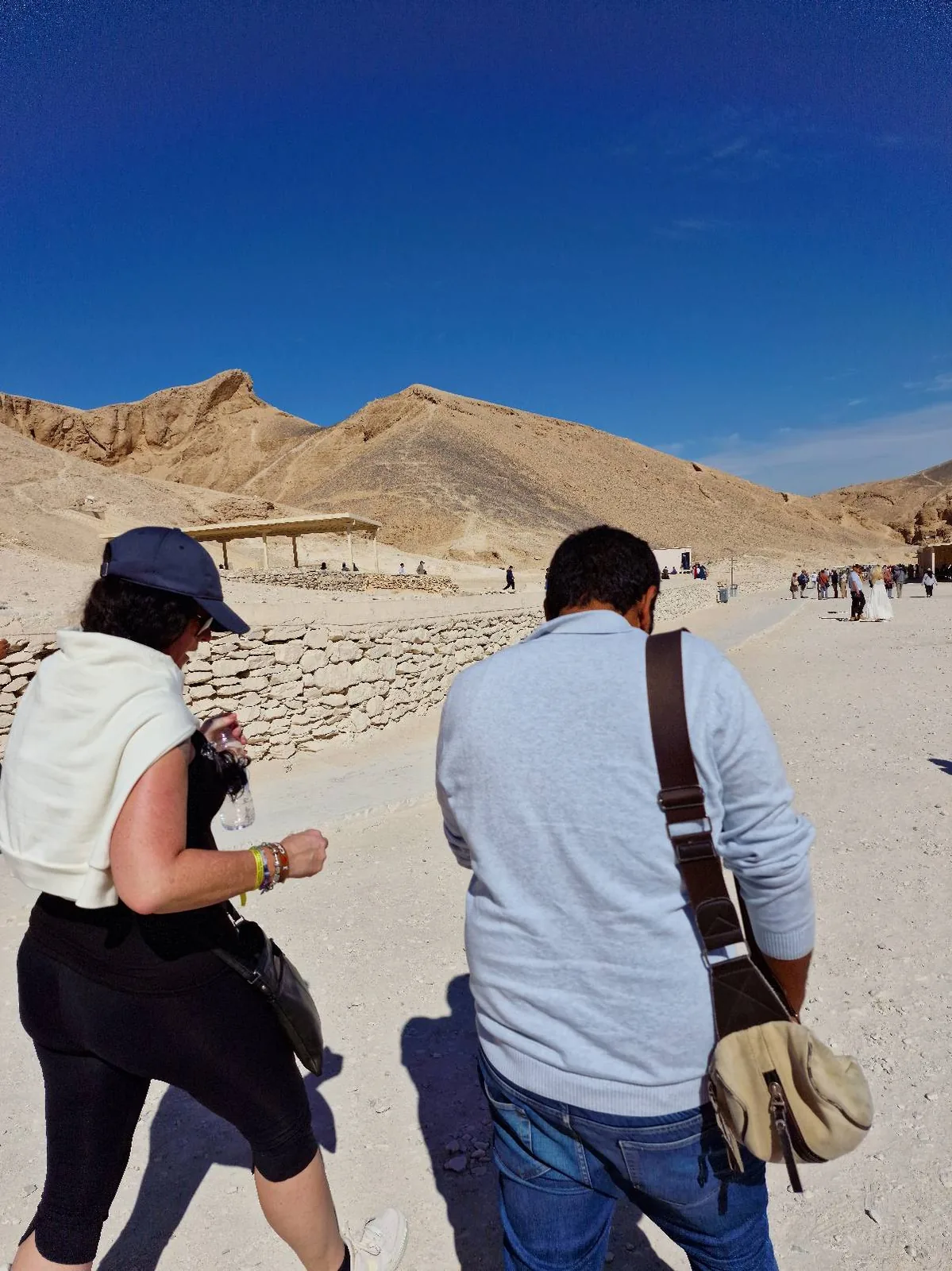 Two people walking in a desert landscape under a clear blue sky. They are on a sandy path, with rocky hills in the background. One person wears a hat and shorts, the other carries a shoulder bag.