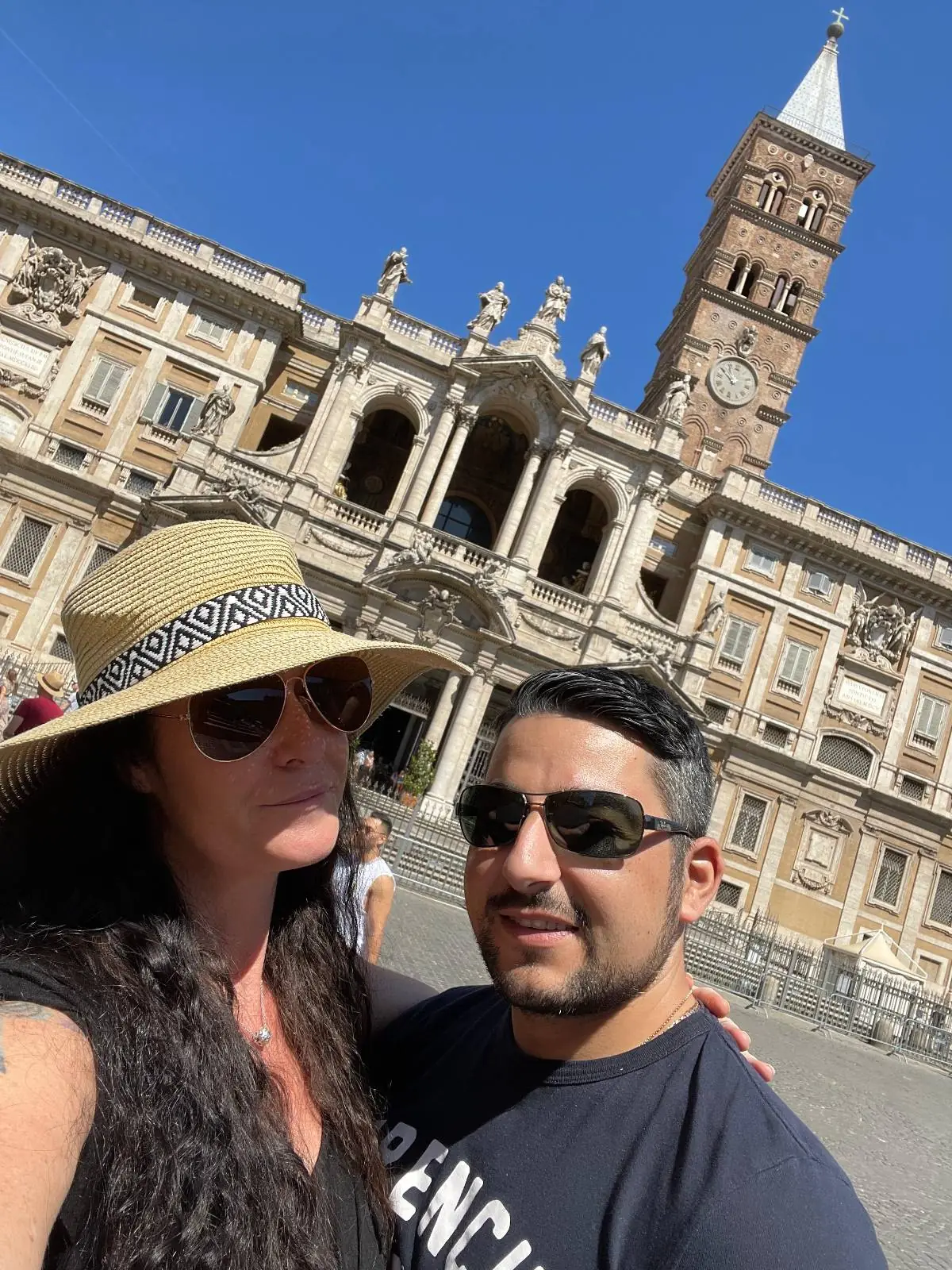 A couple poses for a selfie in front of a historic building with a clock tower and ornate architecture, under a clear blue sky. The woman wears a sun hat and sunglasses; the man wears sunglasses and a dark shirt.