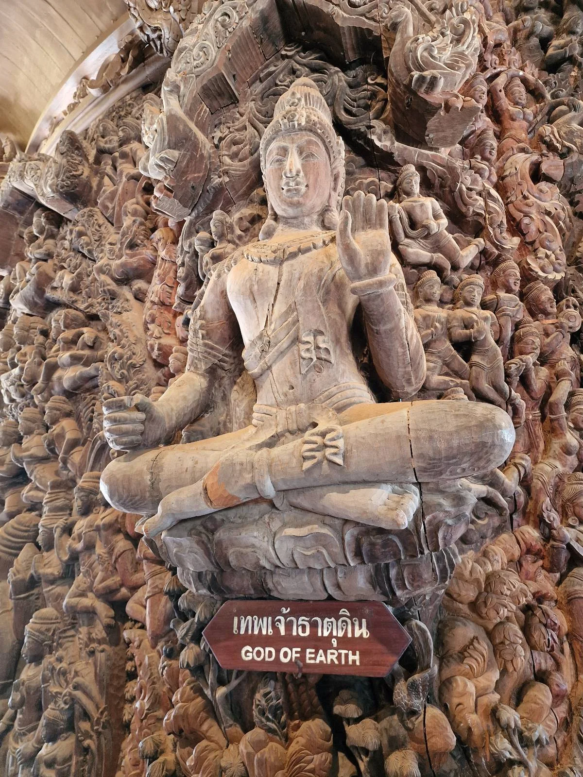 Intricate wooden sculpture depicting a deity seated cross-legged, surrounded by detailed carvings. A sign below reads "God of Earth." The art showcases elaborate craftsmanship and is set against a richly carved background.