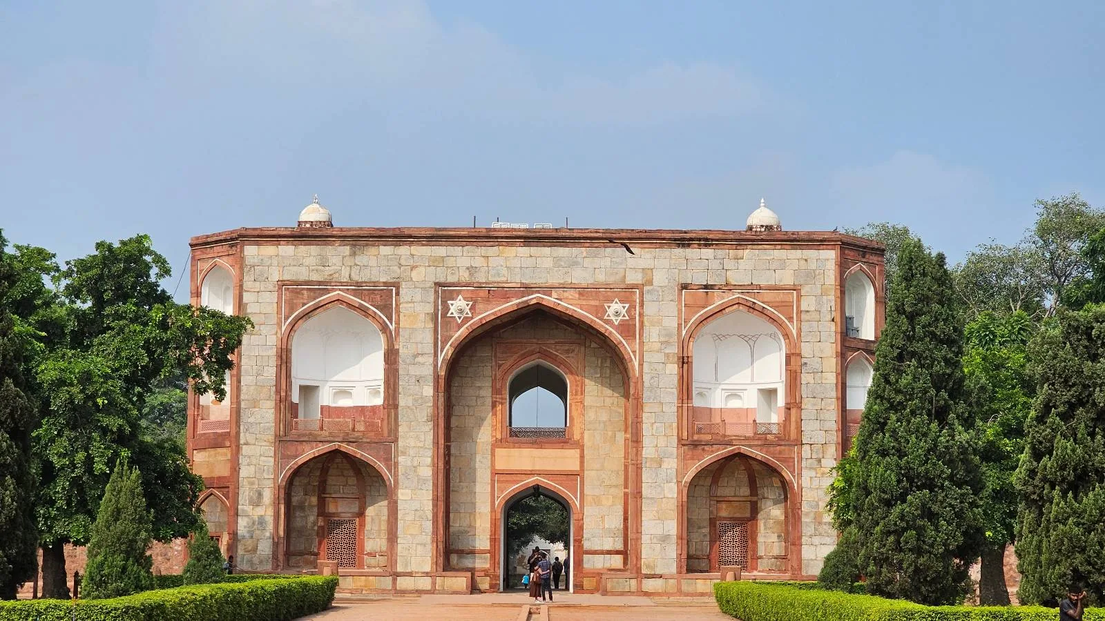 The image shows an arched entrance gate with intricate red and white stonework. It is surrounded by lush green trees and set against a clear blue sky. A pathway leads to the gate, and a few people are walking towards it.