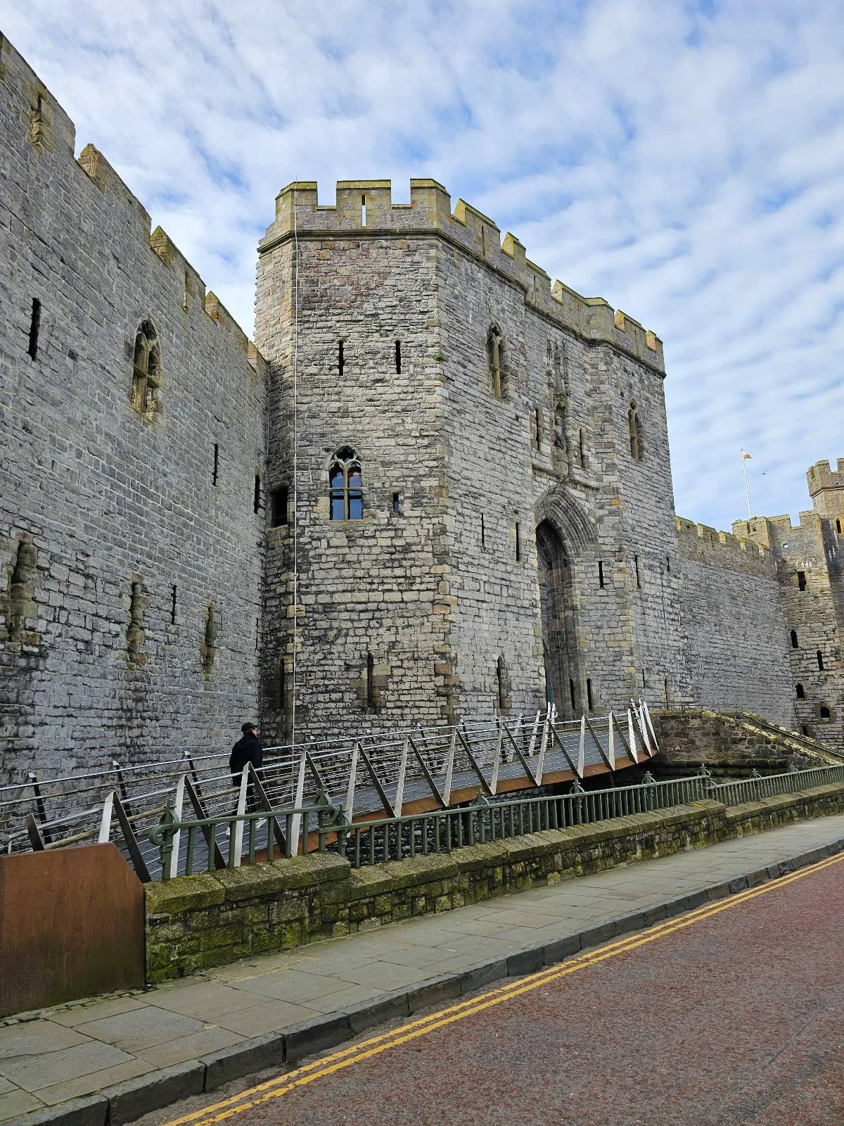 A large stone castle with tall walls and battlements, featuring a central entrance and small windows. A few people walk near the base along a paved path; the sky above is partly cloudy.