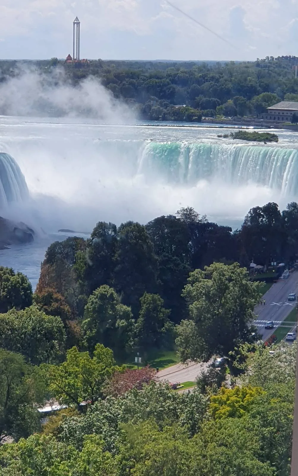 View of Niagara Falls with mist rising above the falling water, seen above treetops and a road.