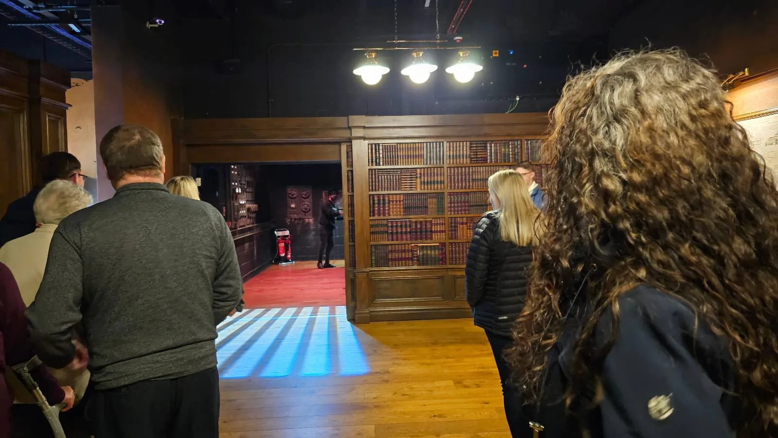 A group of people stands in a dimly lit room with wooden floors and walls lined with bookshelves, looking toward a doorway leading to a brighter room with red carpet and overhead lights.