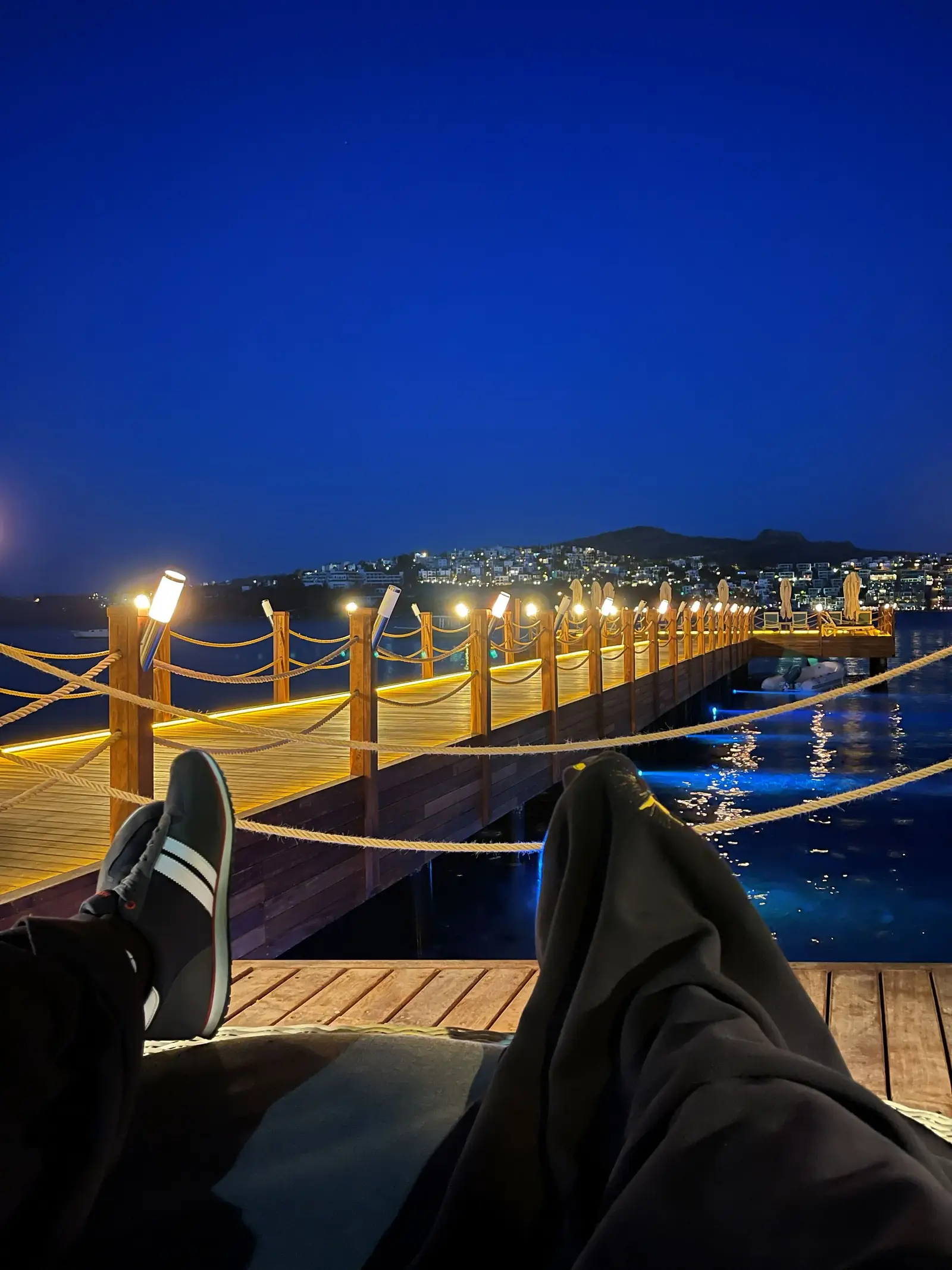 Two people relaxing on a wooden deck at night, overlooking a lit pier and calm water with city lights in the background.