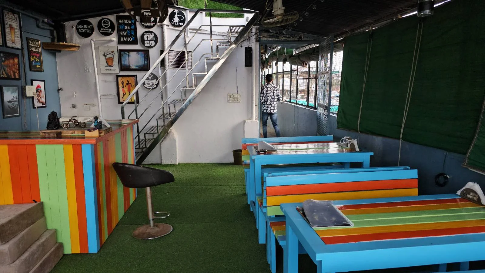 A colorful cafe interior featuring a vibrant, striped counter and matching tables with benches. The floor is covered in green carpet. Framed photos adorn the walls, and a staircase leads up. A person is visible near the stairs.