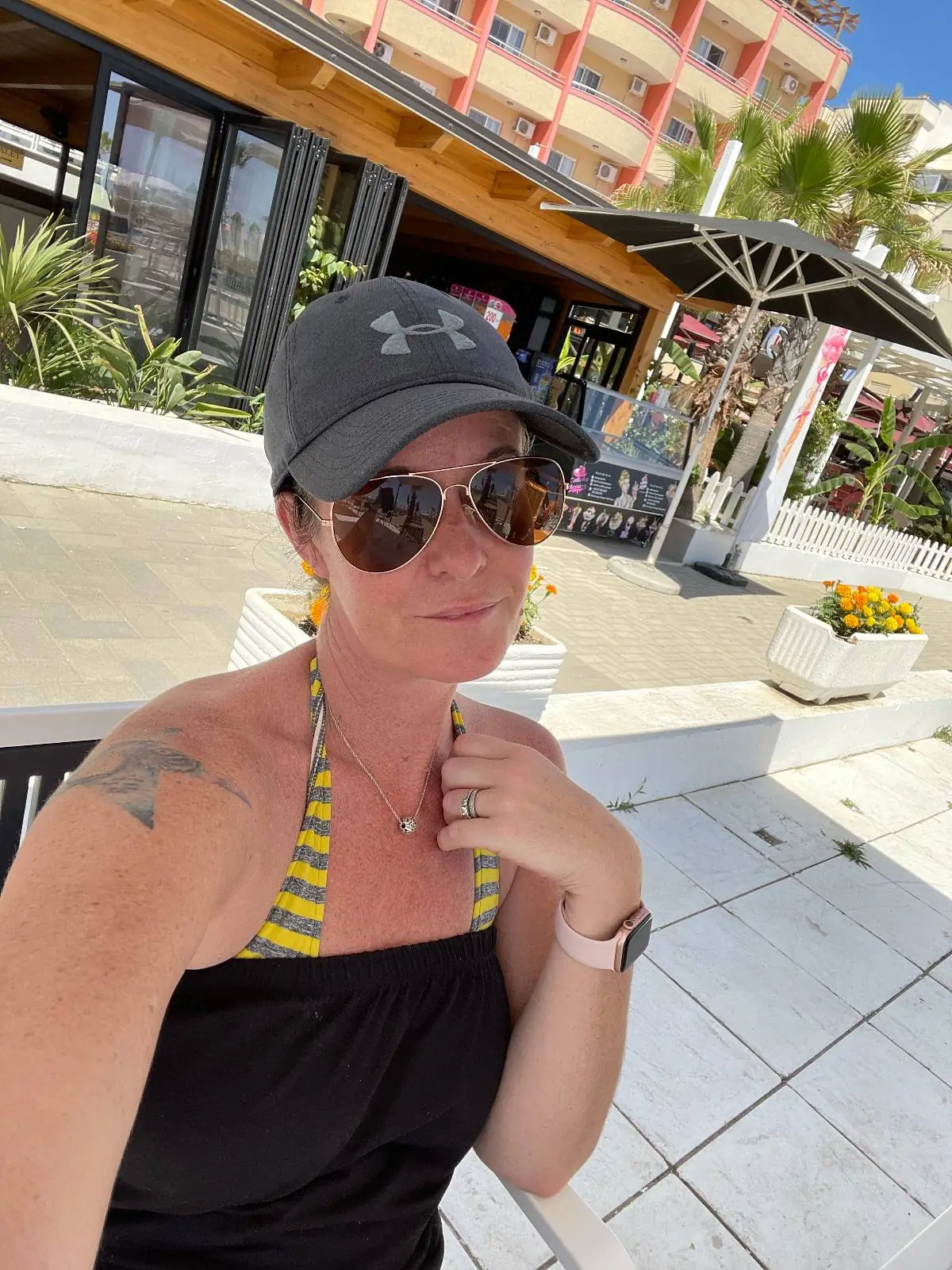 A woman wearing sunglasses, a gray cap, and a black and yellow swimsuit poses for a selfie outdoors. Behind her are potted plants, a building, and sunny weather.