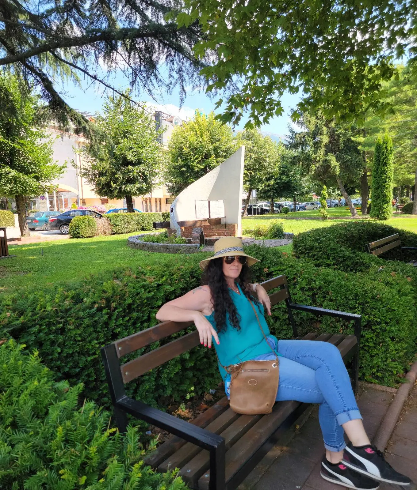 A woman in a straw hat, sunglasses, and teal top sits on a wooden bench in a park. She has a brown crossbody bag and blue jeans. Greenery and a sail-shaped sculpture are visible in the background. It's a sunny day.
