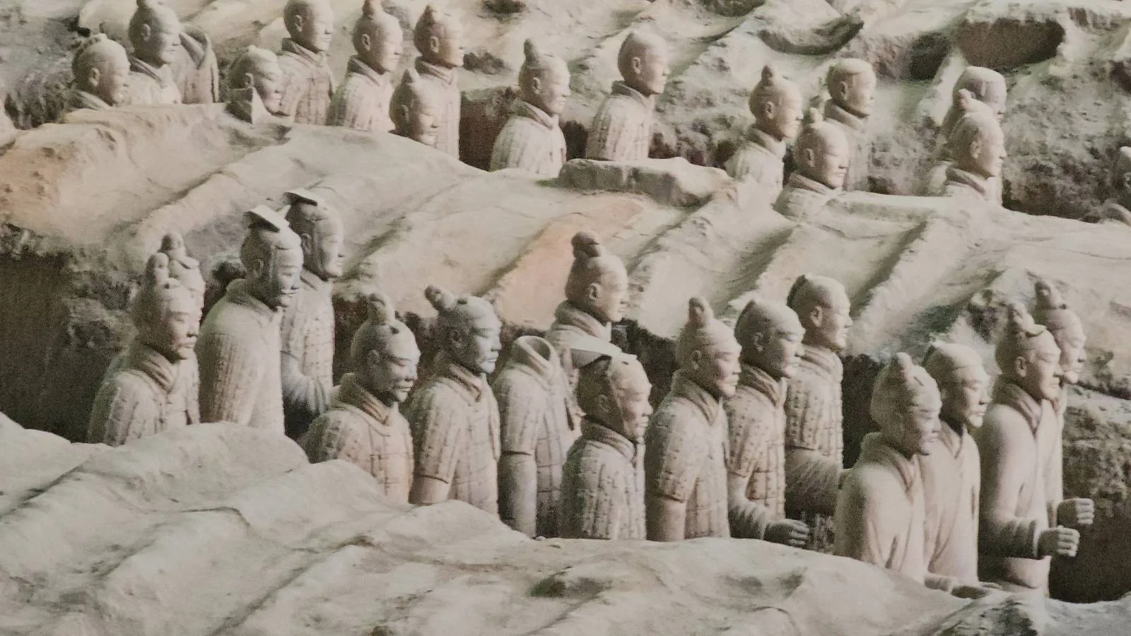 Rows of the Terracotta Army statues, partially buried in earth, stand in an archaeological excavation site in China. The life-sized soldier figures have distinct facial features and are positioned in formation.