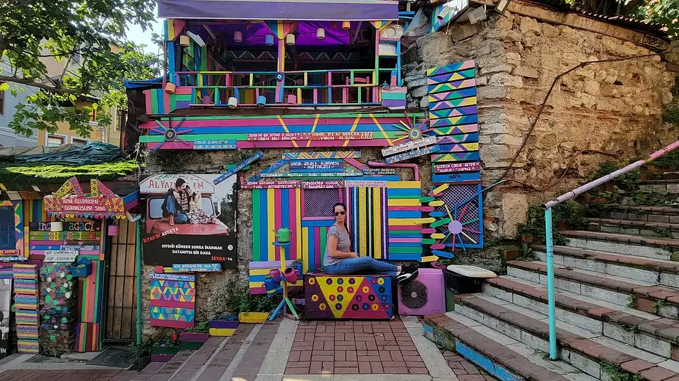 Colorful facade with vibrant patterns and text. A person in sunglasses sits casually on a patterned box. Stone steps at the side.