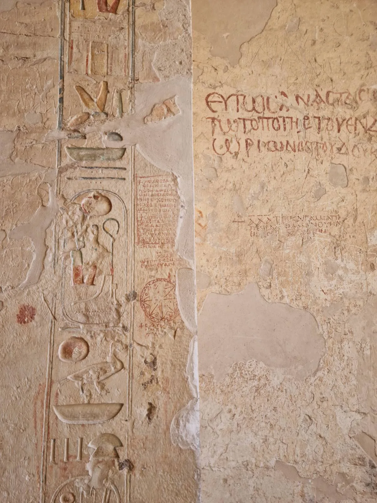 Ancient wall with carved hieroglyphics on the left and Greek inscriptions on the right. The surface is weathered, displaying signs of aging and wear.