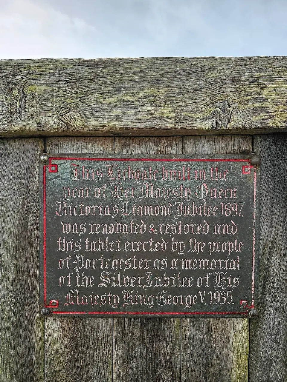 Red plaque mounted on a wooden fence commemorating the restoration of a sluice gate in honor of King George V's silver jubilee, 1935.