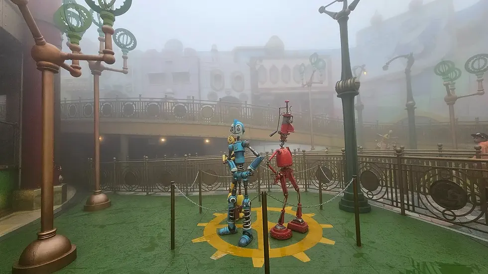 Colorful robots stand on a foggy green platform in a whimsical setting with ornate railings and street lamps. A sign reads "Robot City."