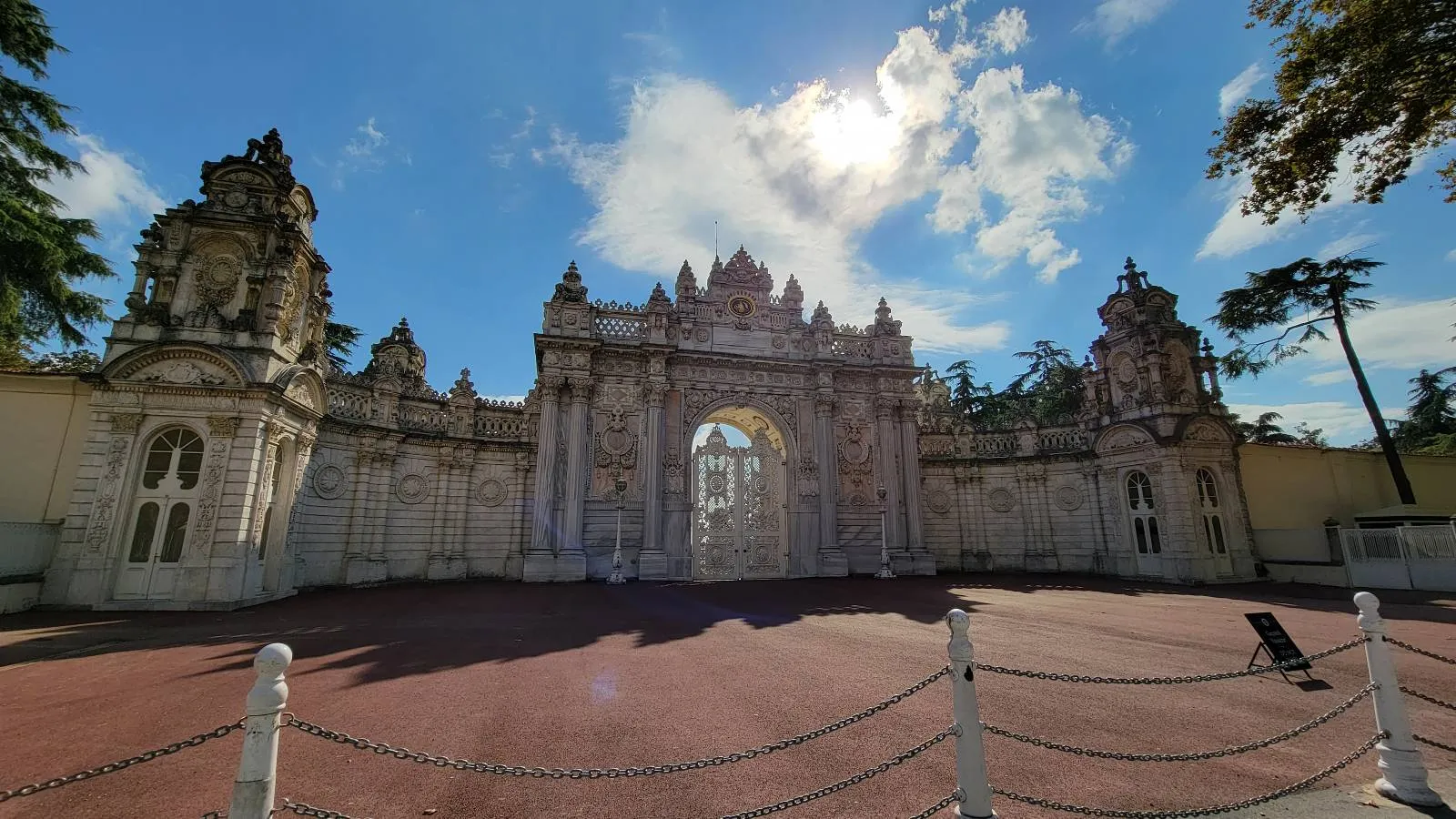 An ornate stone gate with elaborate carvings stands under a blue sky with scattered clouds; sunlight shines above, casting shadows on the brick courtyard in front.