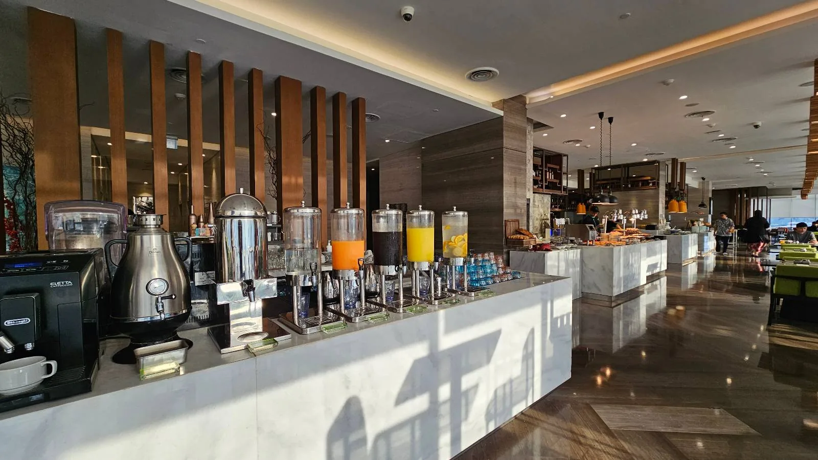 A modern self-serve breakfast buffet with drink dispensers for juice, coffee machines, and various breakfast items on a long, well-lit counter in a spacious dining area.