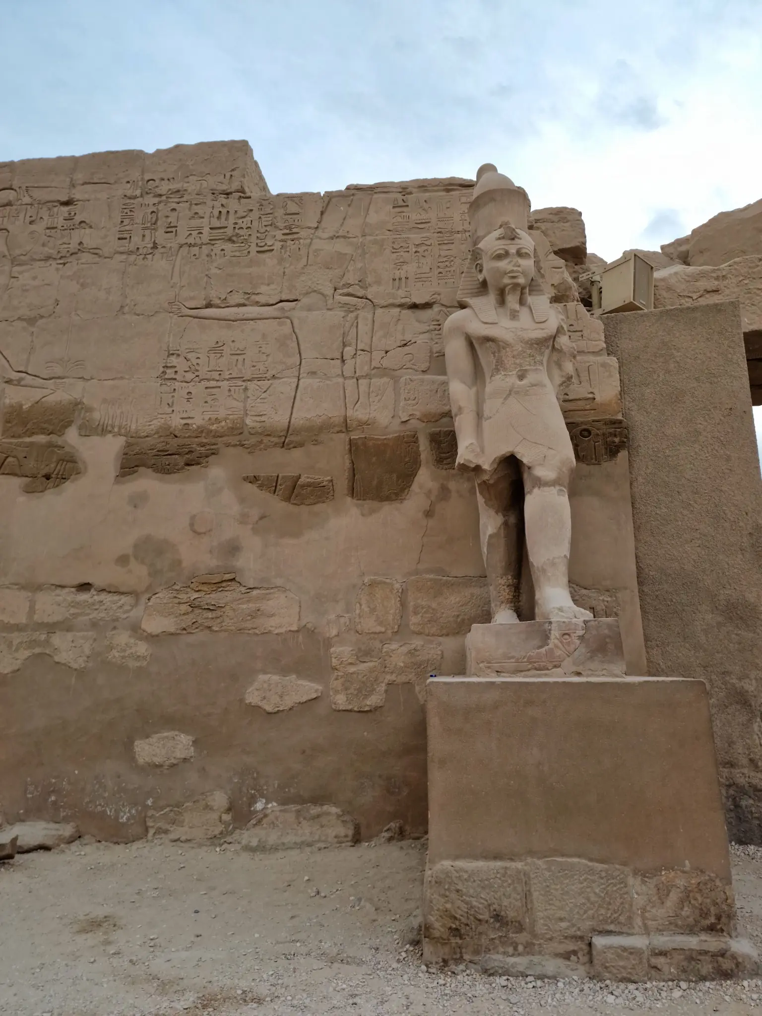 A large stone statue of an ancient figure stands beside a wall with engraved carvings and hieroglyphics, partially damaged and weathered. The figure appears to be wearing a headdress.