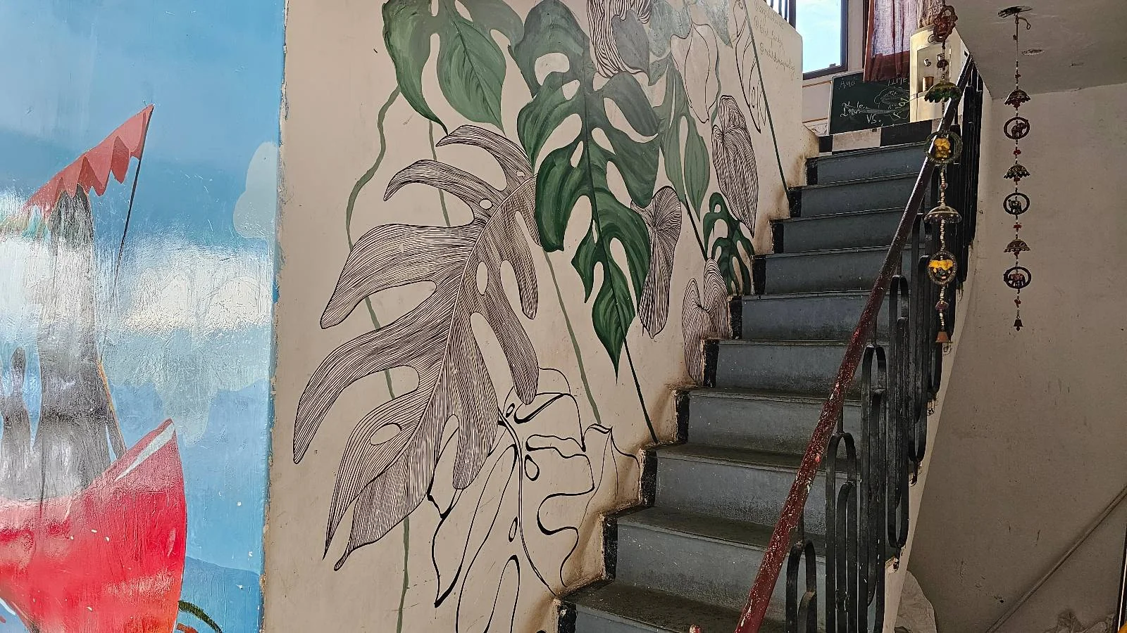 A staircase with a decorative railing beside a wall featuring large, hand-drawn and painted green and brown leaves. To the left, part of a colorful mural with a sky and a red shape is visible.