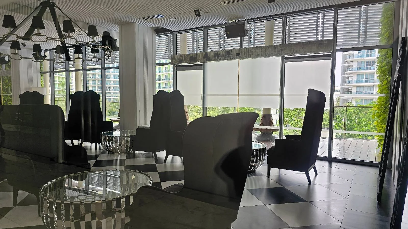 A modern lounge with tall, black high-back chairs, glass tables, and large windows letting in natural light; city buildings and greenery are visible outside.