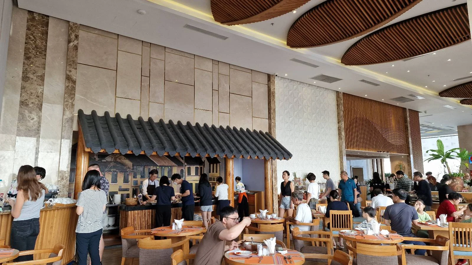 A busy restaurant with patrons sitting at wooden tables and chairs. Some people are in line at a buffet, while others are dining. The interior features high ceilings, warm lighting, and decorative wooden accents on the ceiling.