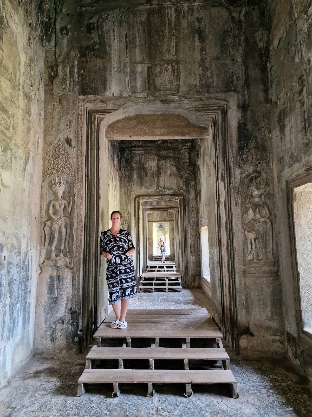 A person in a patterned dress stands on wooden steps inside an ancient stone corridor with multiple doorways and intricate wall carvings. Light enters from windows along the right side.