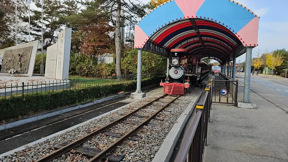 Train on tracks under colorful canopy, surrounded by trees. Nearby, a monument with Korean text and relief sculpture is visible. Calm setting.