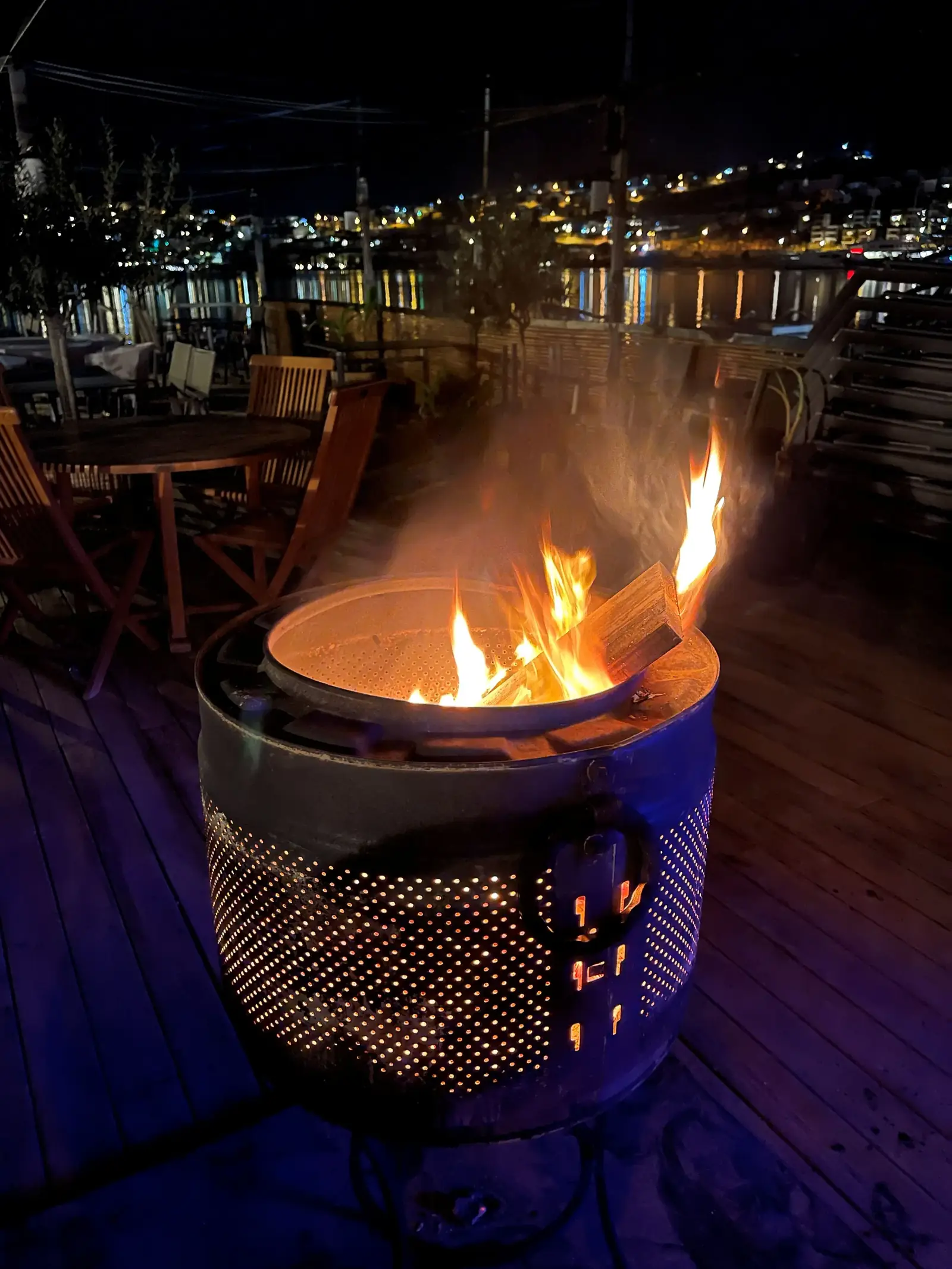 A fire pit on a wooden deck at night, with flames burning inside a metal drum. Outdoor seating and city lights reflecting on the water are visible in the background.