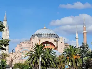 A view of a historic building in Istanbul with a large central dome and four minarets, surrounded by lush green trees and a clear blue sky overhead, capturing the essence of Turkey's architectural beauty.