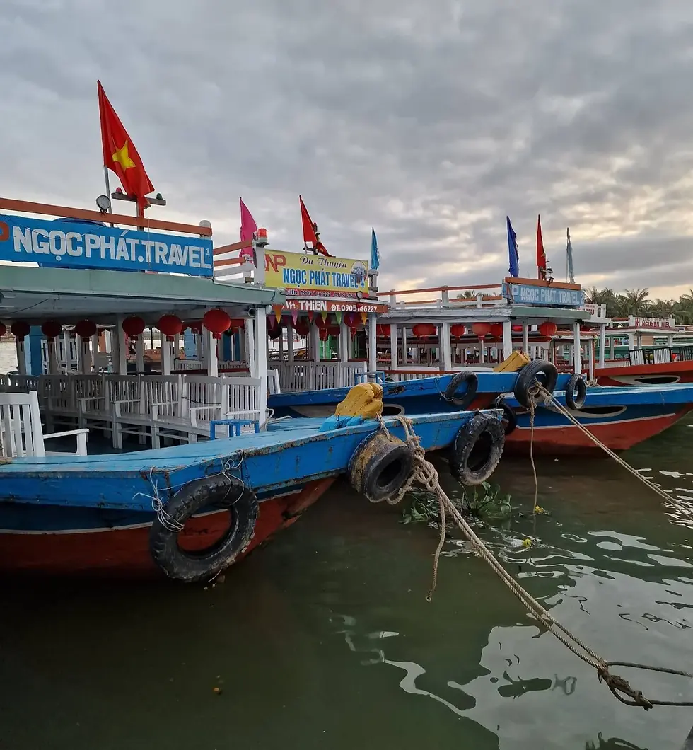 Three colorful boats with flags docked on a calm water body under a cloudy sky.