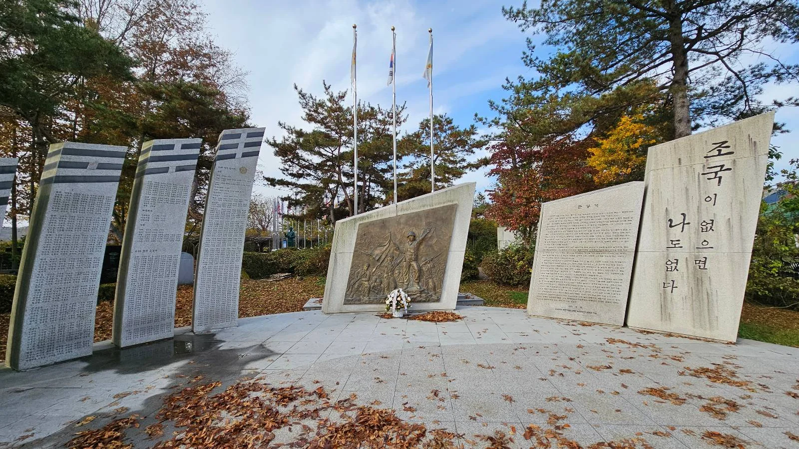 A memorial site with stone monuments featuring engraved text and artwork stands amid trees. Fallen leaves cover the ground, and three flagpoles with flags are seen in the background under a partly cloudy sky.