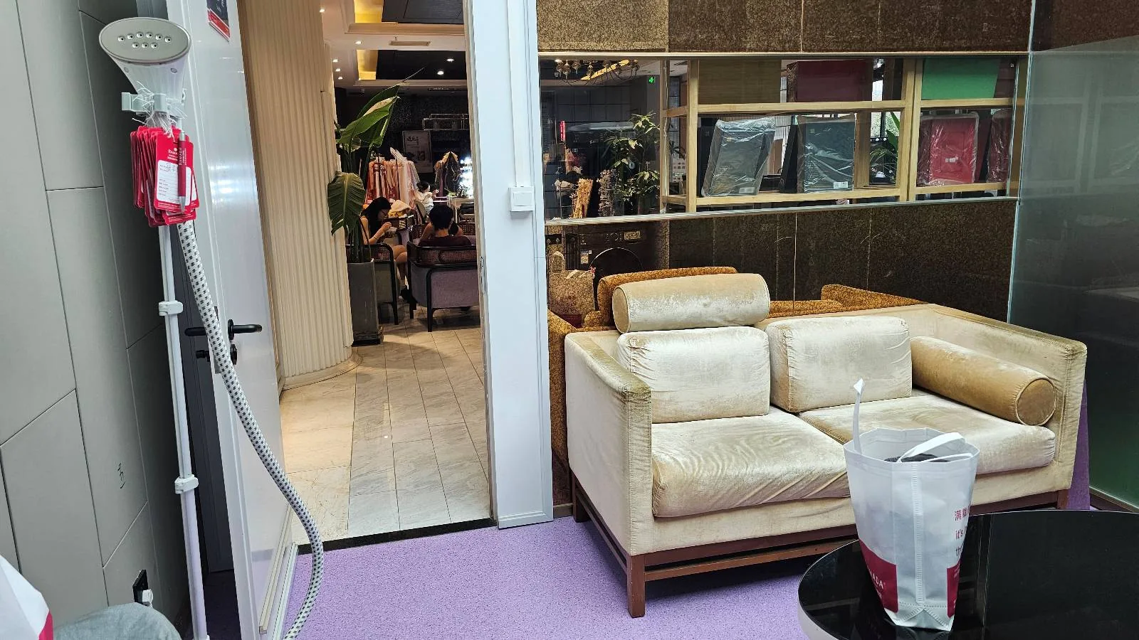 A small room with a beige sofa, glass table with a plastic bag, and a doorway leading to a busy area with people and bright lights. A garment steamer stands near the door on a purple floor.